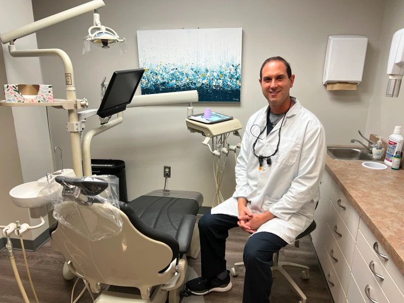 Dr. Adam Klein sitting beside a dental chair in a treatment room at Monroe Dental Group